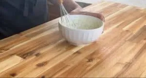 Person whisking creamy mixture in a white bowl on a wooden kitchen countertop.