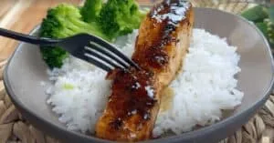 Grilled salmon with glossy glaze served on white rice with a side of fresh broccoli in a beige bowl.