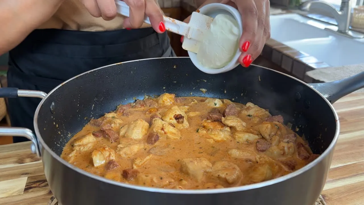 Person adding cream cheese to a simmering chicken and chorizo dish in a black skillet on a wooden countertop.