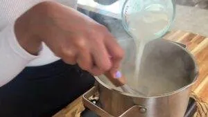 Person stirring steaming pot while pouring liquid from measuring cup on wooden table. Cooking process close-up.