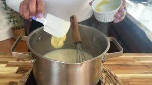 Woman stirring homemade soup with a whisk in a pot, adding a yellow ingredient using a spatula.
