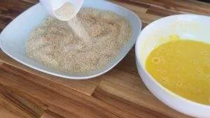 Breadcrumbs being poured onto a plate next to a bowl of beaten eggs for coating food before frying.