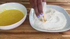 Hand adding spices to flour beside a bowl of beaten eggs on a wooden countertop.