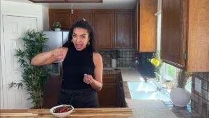 Woman excitedly seasoning a dish in a cozy kitchen with wood cabinets and a sunlit counter.