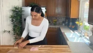 Woman preparing a ham and cheese sandwich in a cozy kitchen with wooden cabinets and plants.