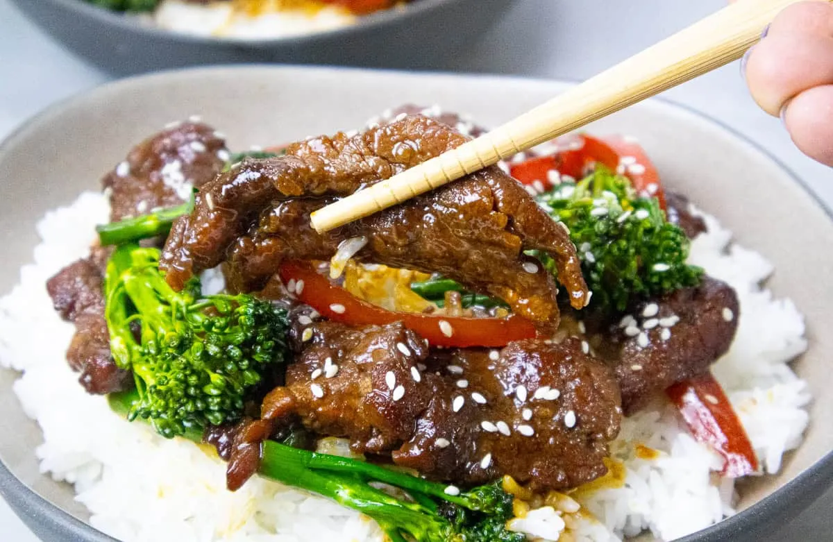 Beef stir-fry with broccoli, red peppers, and sesame seeds on rice, held with chopsticks. Asian cuisine dish.