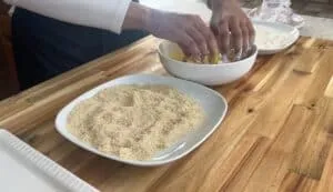 Person breading food with egg wash and breadcrumbs on a wooden table.