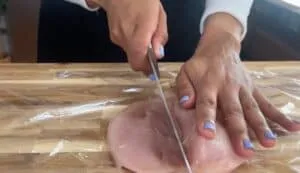 Person slicing raw chicken breast on a wooden cutting board for meal preparation.
