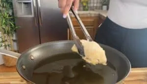 Person frying breaded chicken in a pan using tongs in a modern kitchen.