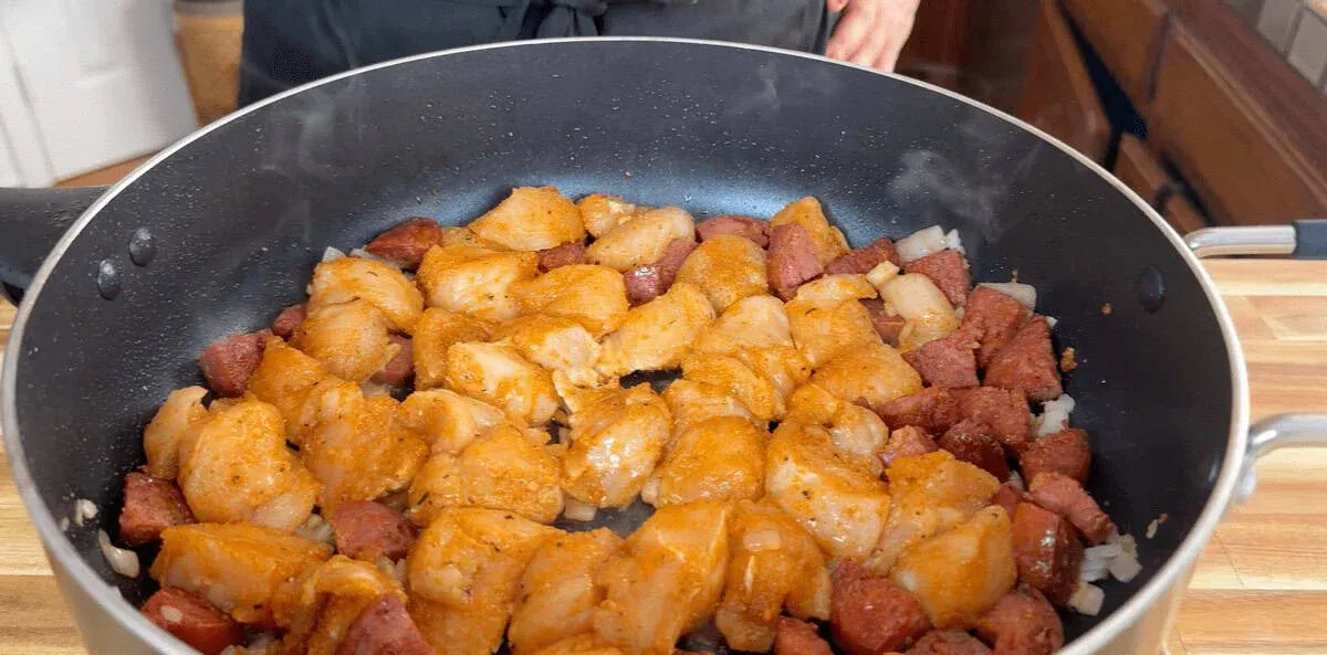 Cooking seasoned chicken and sausage in a skillet on a wooden countertop.