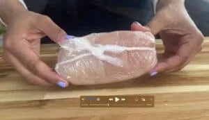 Hands wrapping chicken breast in plastic wrap for sous vide cooking.