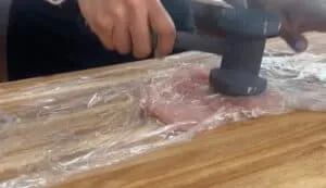 Pounding raw chicken with meat tenderizer on wooden board, covered in plastic wrap.