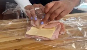 Hands wrapping ham and cheese slices in plastic wrap on a wooden cutting board.