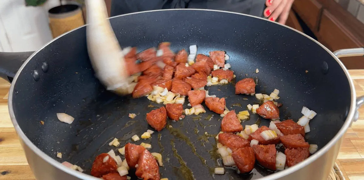 Cooking sliced sausage with onions in a pan, stirred with a wooden spoon on a stovetop.