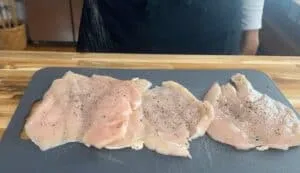 Raw chicken fillets being seasoned with salt and pepper on a cutting board in a kitchen setting.