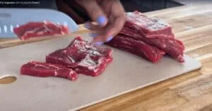 Person slicing fresh beef steak on a wooden chopping board, preparing for a stir fry meal.