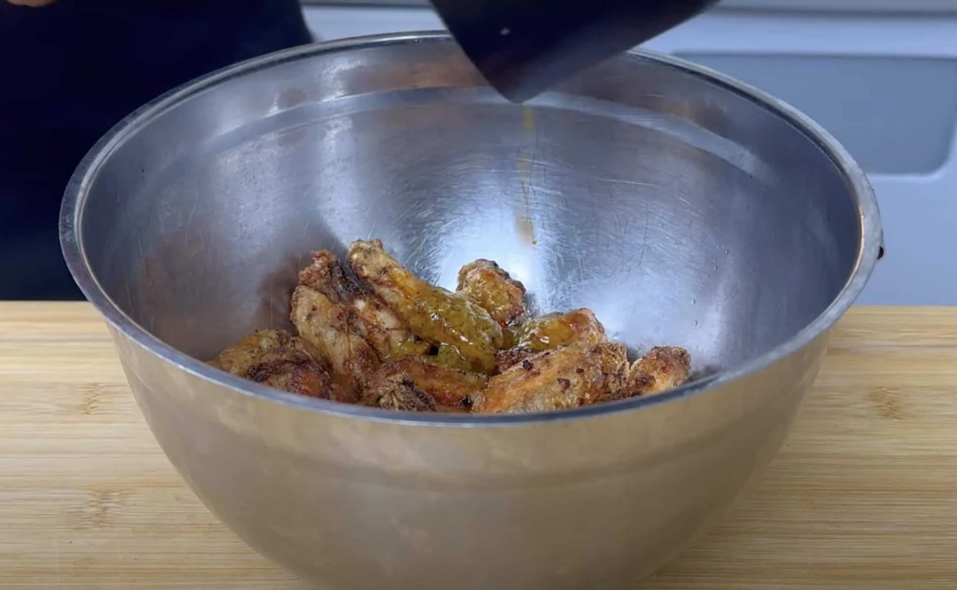 Pouring sauce over crispy chicken wings in a stainless steel bowl on a wooden surface.