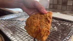 Hand holding a crispy fried chicken fillet over a cooling rack in a kitchen setting.
