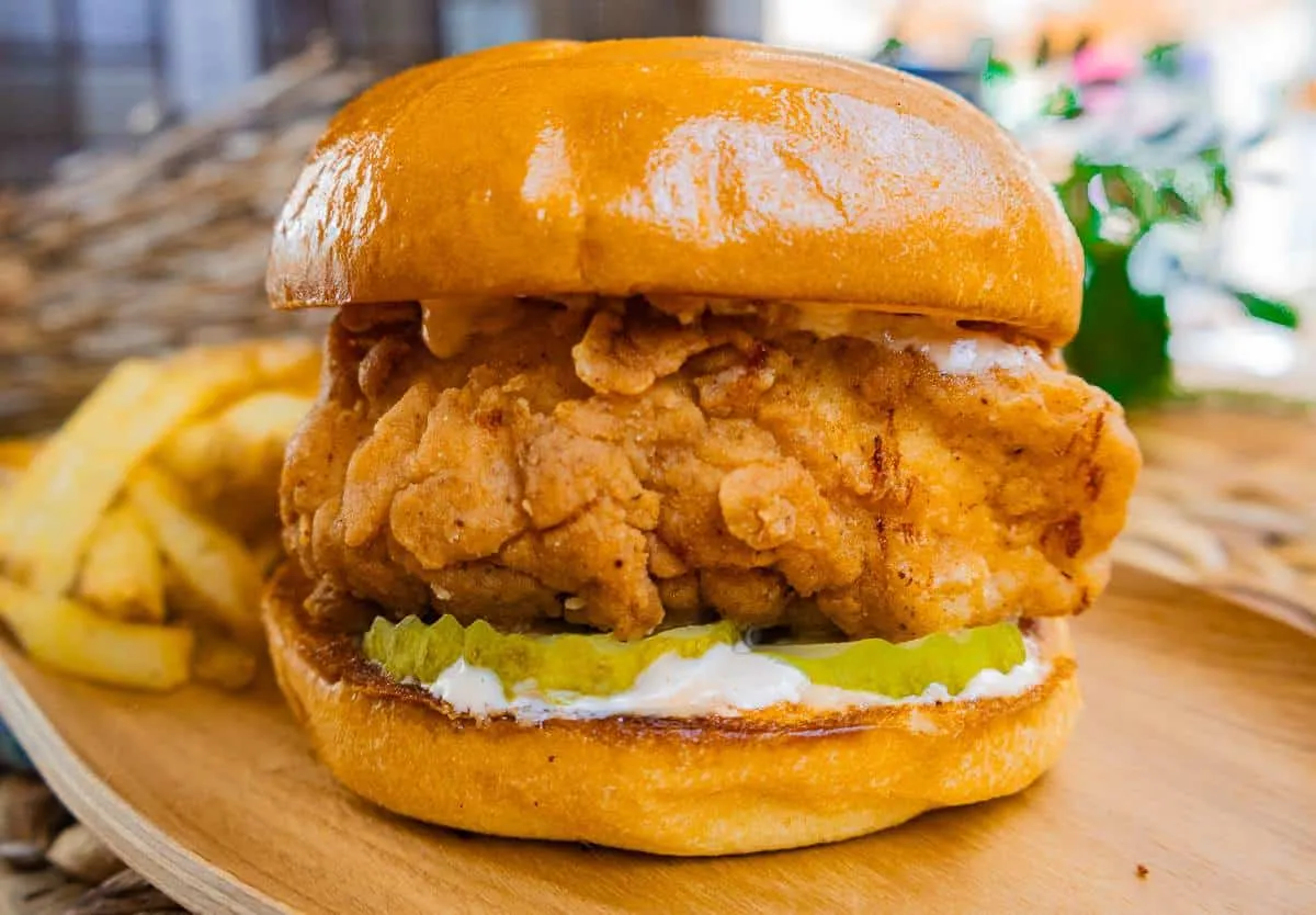 Crispy fried chicken sandwich with pickles on a brioche bun, served with fries on a wooden plate.