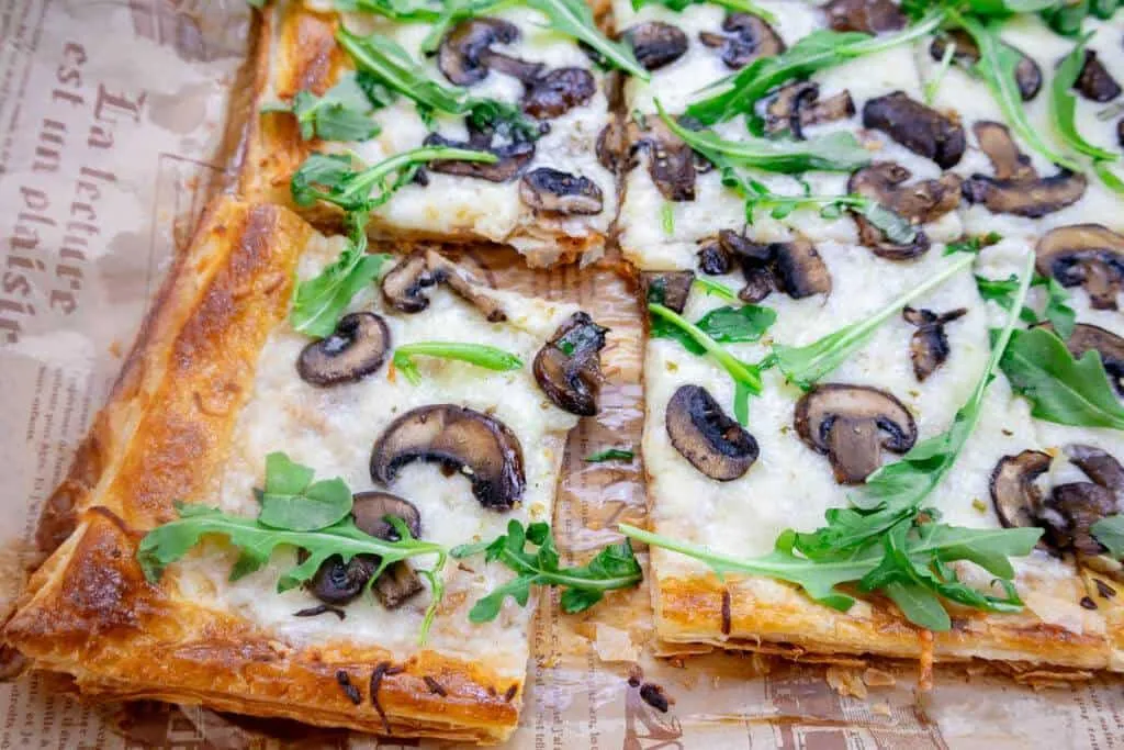 Crispy puff pastry topped with mushrooms, arugula, and cheese on parchment paper.