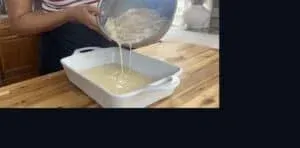 Person pouring batter-covered chicken into a white baking dish on a wooden countertop.