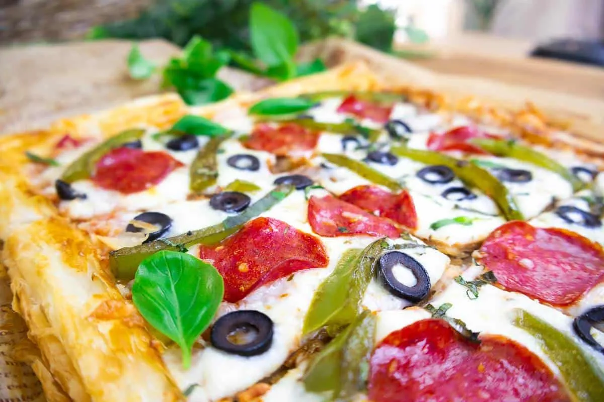 Delicious homemade pizza with pepperoni, olives, green peppers, and basil leaves on a wooden board.