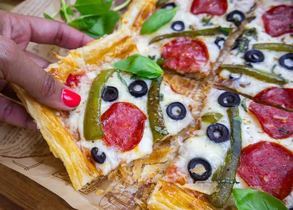 Pizza slice with pepperoni, olives, and green peppers being picked up, on a wooden table garnished with fresh basil.