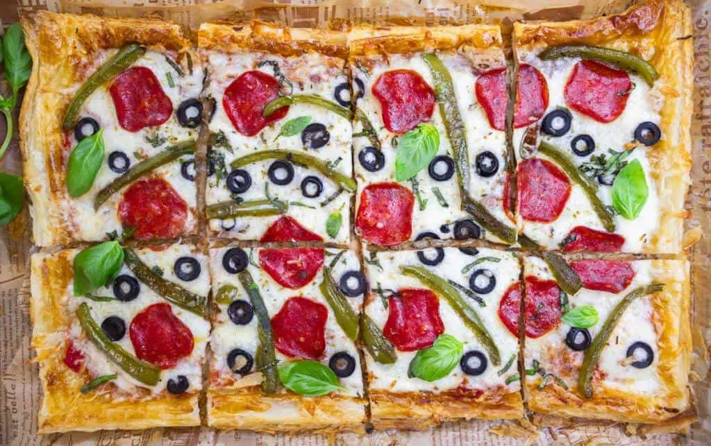 Sliced pizza topped with pepperoni, black olives, green peppers, basil leaves on parchment paper.
