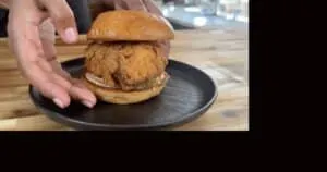 Close-up of a homemade fried chicken sandwich with pickles on a bun, served on a black plate.