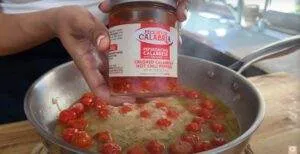 Jar of Calabrian chili peppers held over a skillet with cherry tomatoes cooking.