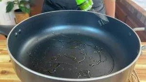 Olive oil being poured into a nonstick frying pan on a wooden countertop in a kitchen setting.