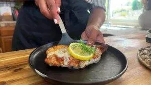 Grilled chicken with creamy sauce, lemon, and dill garnish on a black plate, served by a chef on a wooden counter.
