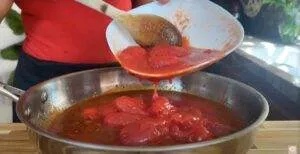 Pouring canned tomatoes into a skillet for cooking sauce, with a wooden spoon stirring gently.