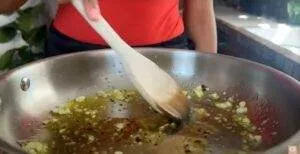 Cooking chopped garlic in oil with a wooden spoon in a large stainless steel pan.