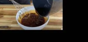 Mixing homemade barbecue sauce in a white bowl with a whisk on a wooden table.