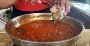 Person adding canned tuna to a tomato-based sauce in a pan on a stove.