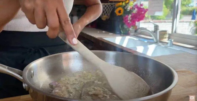 Person cooking onions in a pan with a wooden spoon in a kitchen with flowers on the counter.