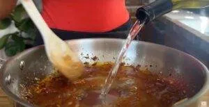 Pouring liquid into a pan to make a sauce, with a wooden spoon stirring, in a kitchen setting.