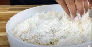 Hand mixing flour in a white bowl for baking preparation on a wooden surface.