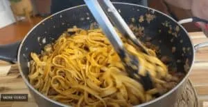 Cooking fettuccine pasta with meat sauce in a pan using tongs on a wooden counter. Perfect for Italian cuisine lovers.