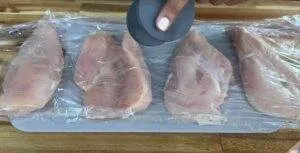 Four chicken breasts covered in plastic wrap being tenderized on a cutting board with a kitchen tool.