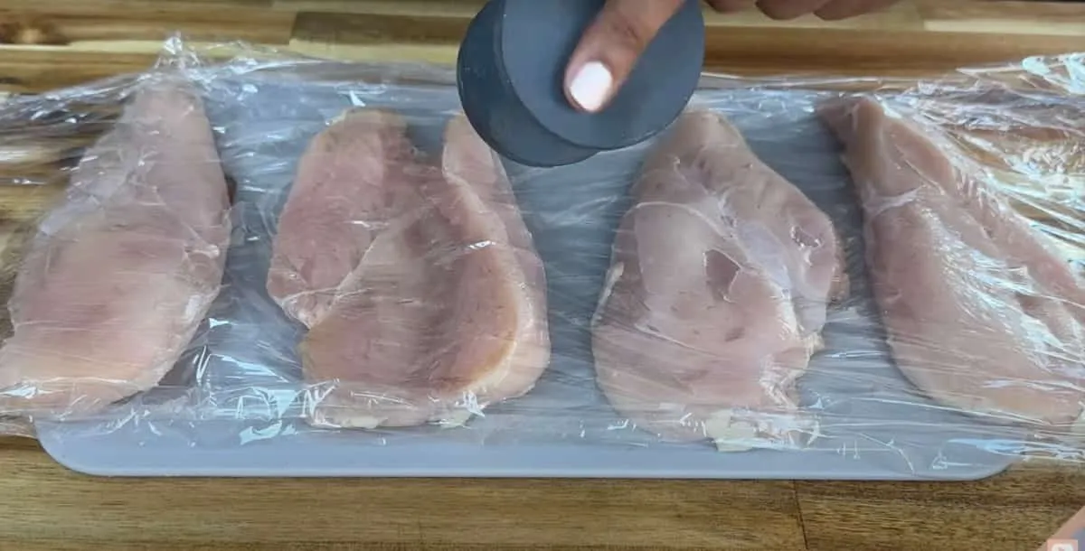 Four chicken breasts covered in plastic wrap being tenderized on a cutting board with a kitchen tool.