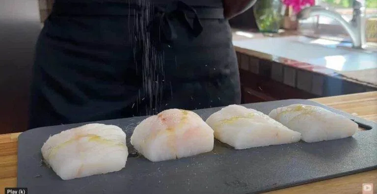 Person seasoning four pieces of raw fish on a cutting board in a kitchen setting.