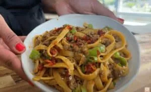 Close-up of spicy beef noodle dish with green peppers and chili flakes in a bowl, held by a person with red nails.