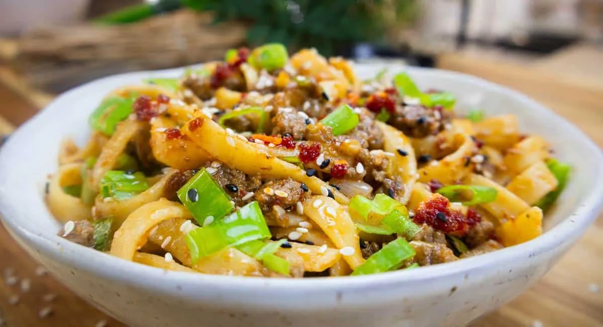 Spicy stir-fried noodles with beef, green onions, and sesame seeds in a white bowl, served on a wooden table.