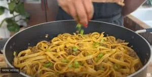 Person seasoning a pan of fettuccine with green onions and meat.
