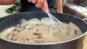 Person whisking creamy mushroom sauce in a black pan.