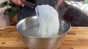Tongs lifting cooked rice noodles from a steaming metal bowl on a wooden countertop.
