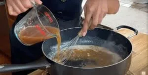 Pouring brown liquid from a measuring cup into a saucepan on a stove, while stirring with a whisk.