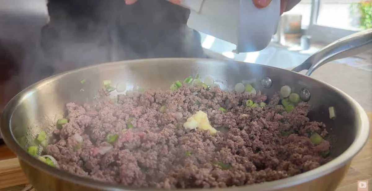 Cooking ground beef with scallions and spices in a skillet. Hands adding seasoning for a flavorful dish.